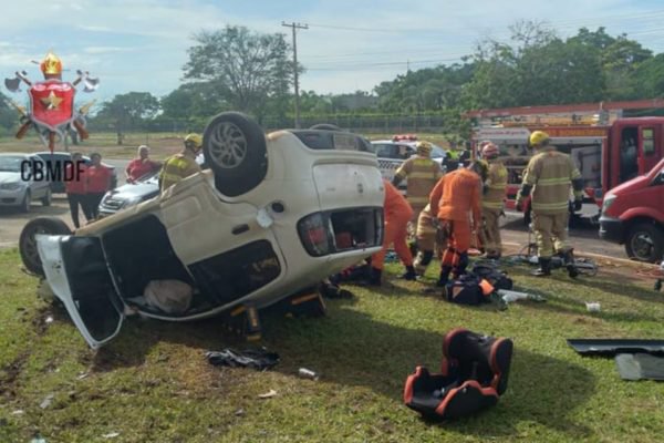 Imagem colorida com carro branco capotado perto da catedral de Brasília. Bombeiros com roupa laranja atuam no resgate