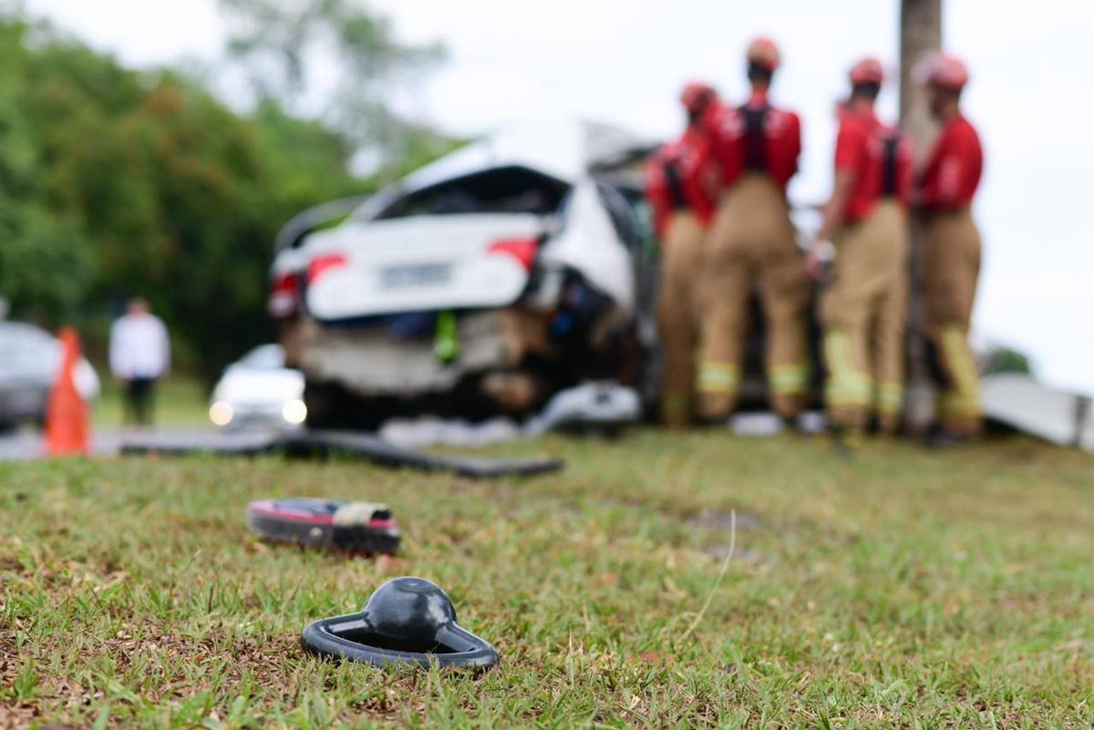 Imagem desfocada de carro branco destruído após acidente, ambulância e bombeiros do lado