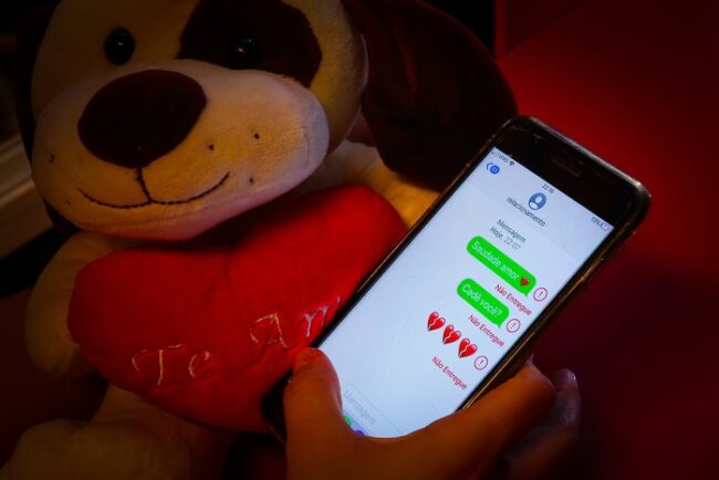 Imagem colorida de um urso, um coração vermelho, escrito 'Te Amo', uma mão segurando um celular com mensagens referentes a relacionamento