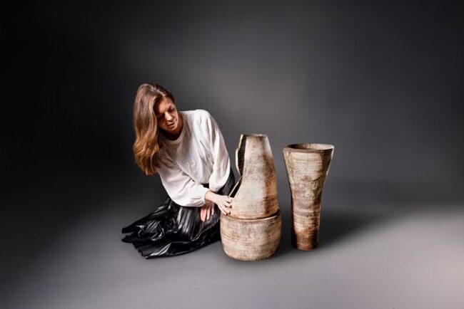 Foto colorida mostra uma mulher ao lado de um vaso quebrado