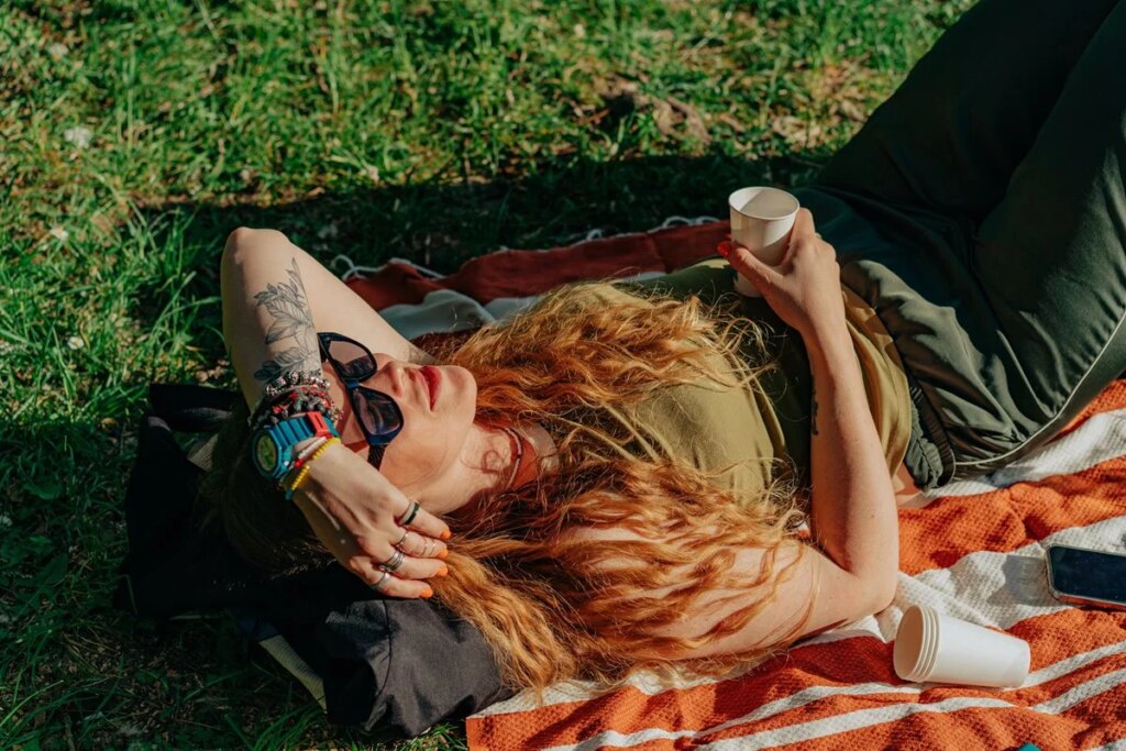 Mulher ruiva deitada no gramado, aproveitando o dia ensolarado e a natureza. Conceito de diversão ao ar livre. Metrópoles