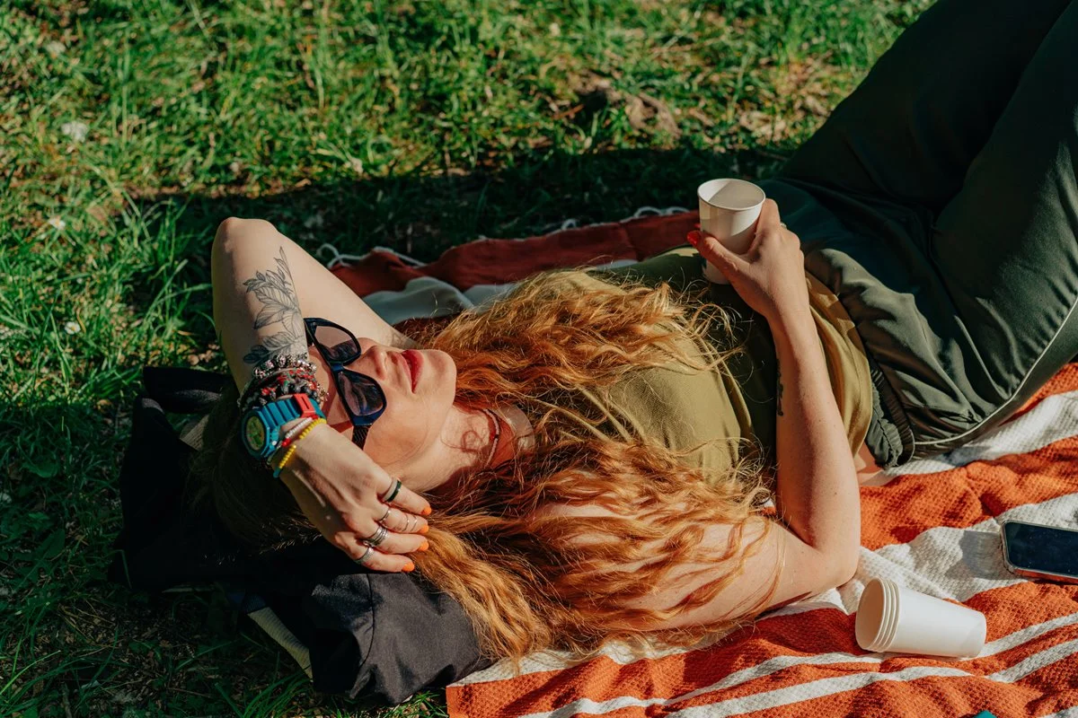 Mulher ruiva deitada no gramado, aproveitando o dia ensolarado e a natureza. Conceito de diversão ao ar livre. Metrópoles